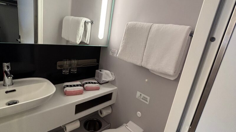 Bathroom interior of a Virgin Voyages cruise ship cabin, featuring a modern sink, toilet, large mirror with integrated lighting, and neatly hung towels.