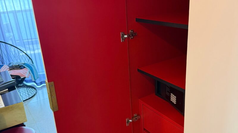 Red closet with open door in a Virgin Voyages cruise ship cabin, revealing shelves, drawers, and a safe. Background includes a balcony door with partial view of a chair.