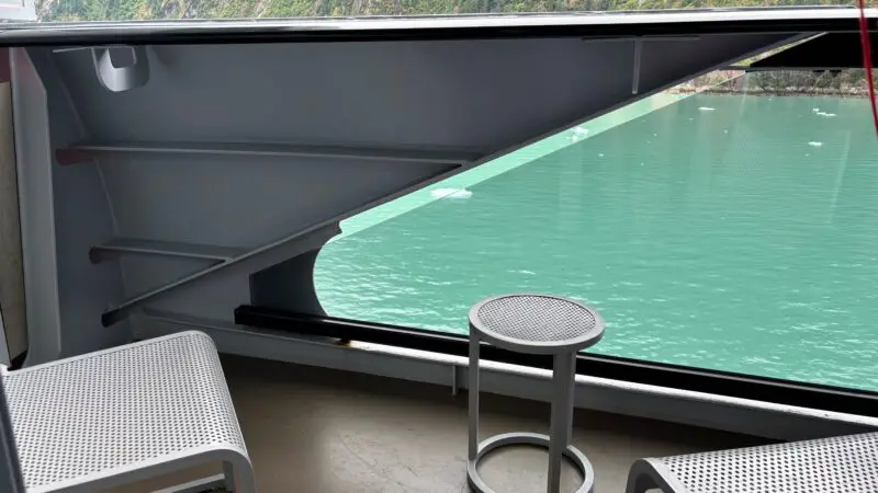 A Virgin Voyages cruise ship balcony with two perforated metal chairs, a small round table, and a view of turquoise water and lush green cliffs in the background.