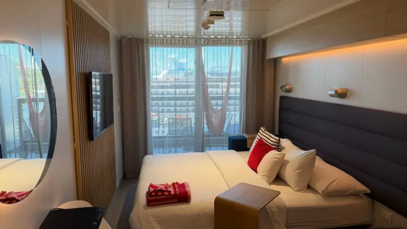 A Virgin Voyages cruise ship cabin featuring a neatly made bed with red and striped cushions, a wall-mounted TV, a round mirror, and a balcony with sheer curtains and a hammock outside.
