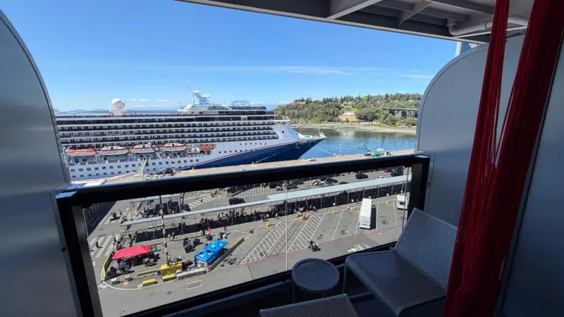 Virgin Voyages cruise ship stateroom balcony with seating and a red hammock, overlooking a docked cruise ship and a distant landscape.