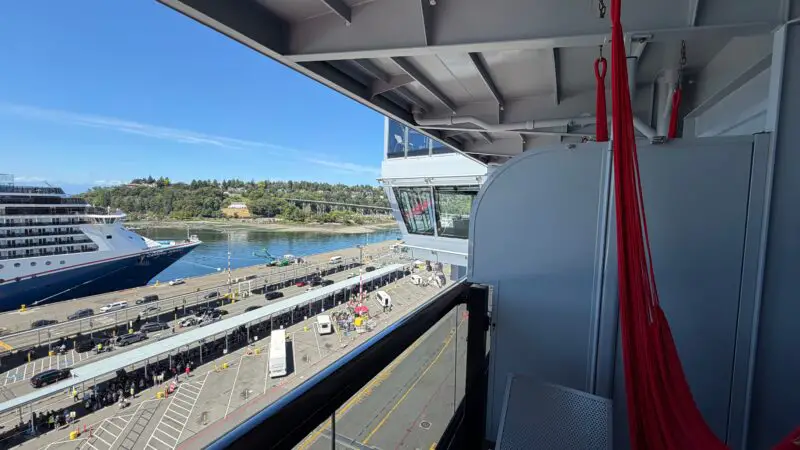 Balcony of a Virgin Voyages cruise ship cabin with a red hammock and chair, overlooking a dock with parked vehicles and another cruise ship in the water.