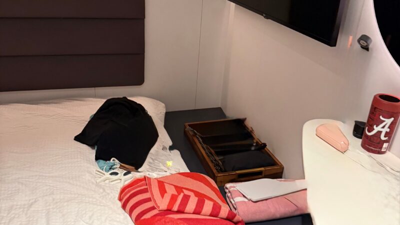 Interior view of a Virgin Voyages cruise ship cabin featuring a bed with white linens and a red-striped blanket. A flat-screen TV is mounted on the wall, and a small table holds personal items.