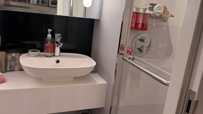 Bathroom of a Virgin Voyages cruise ship cabin, featuring a sink, mirror, toiletries, and shower with glass door.