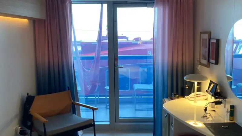 Virgin Voyages cruise ship cabin with a balcony. Features include a bed, chair, desk with lamp, and a window leading to an outside deck. Pink and blue curtains partially cover the glass door.