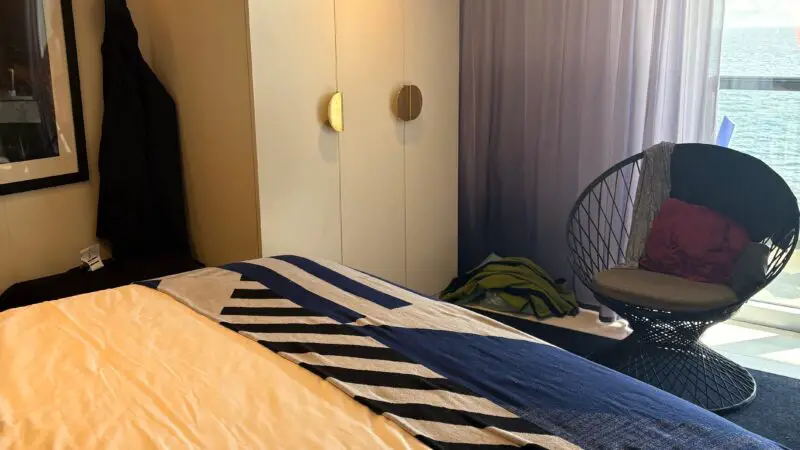 Interior of a Virgin Voyages cruise ship cabin, featuring a bed with a striped blanket, a wardrobe, a chair next to a window with curtains, and a glimpse of the ocean outside.