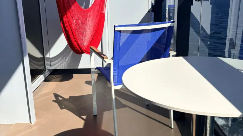 A Virgin Voyages cruise ship balcony featuring a red hammock, a blue chair, and a white round table, with ocean views in the background.