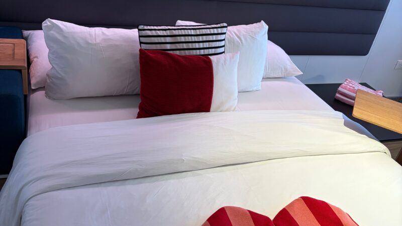 Virgin Voyages cruise ship cabin with a bed, featuring decorative pillows and a folded heart-shaped towel on the duvet.