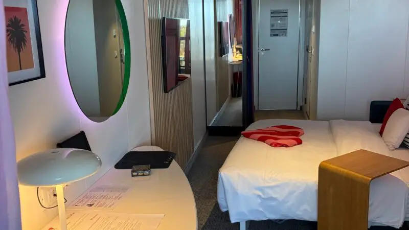 Virgin Voyages cruise ship cabin with a bed, a side table, a wall-mounted TV, a round mirror, and a desk with a lamp and papers.