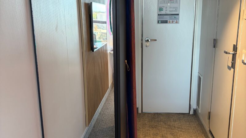 Virgin Voyages cruise ship cabin hallway featuring a full-length mirror, a wall-mounted TV, and a closed door with an evacuation plan.