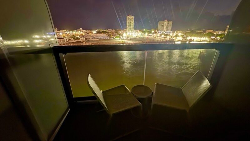 Virgin Voyages cruise ship cabin balcony at night, featuring two chairs and a small table, overlooking a cityscape with illuminated buildings and waterfront.