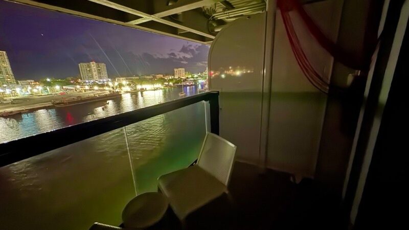 A Virgin Voyages cruise ship cabin balcony at night, with chairs and a small table overlooking a lit-up waterfront cityscape.