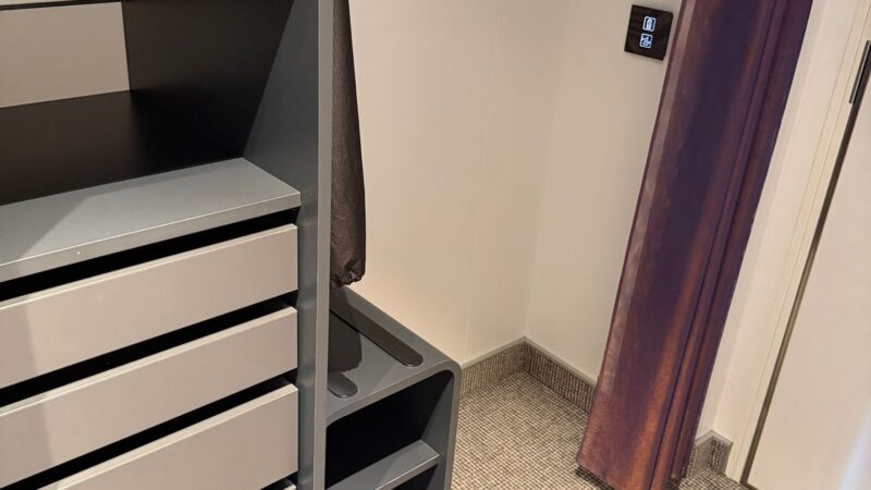 Interior view of a Virgin Voyages cruise ship cabin featuring shelves, a safe, and a wall control panel next to a curtain.