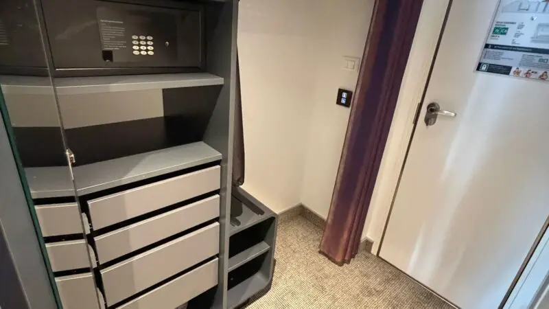 A Virgin Voyages cruise ship cabin closet, featuring shelves with a safe, drawers, metal hangers, and a curtain on a rod. Carpeted floor and a white door with an evacuation plan visible.