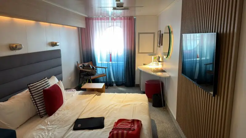 Virgin Voyages cruise ship cabin featuring a neatly made bed with striped pillows, a window with blue and pink curtains, a wall-mounted TV, a desk with a laptop, and a chair.