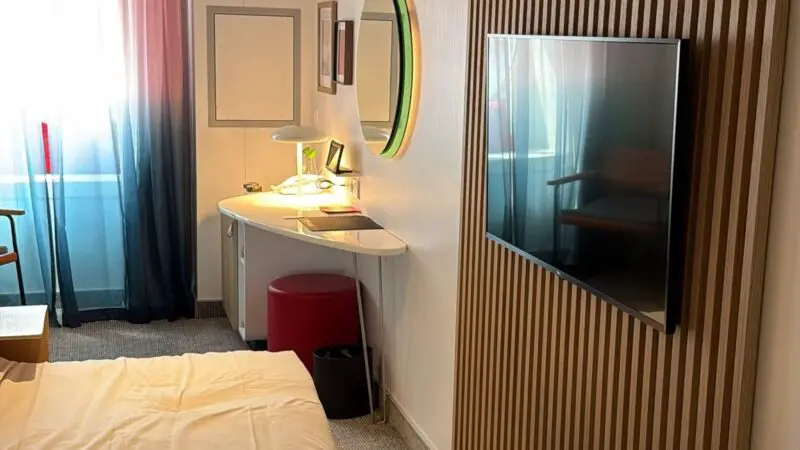 Virgin Voyages cruise ship cabin featuring a bed with red-striped towel, wall-mounted TV, desk with mirror, and window with sheer curtains.