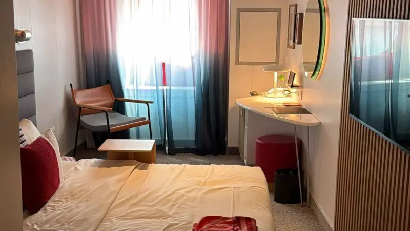 Virgin Voyages cruise ship cabin featuring a bed with red pillows, a small table, a chair, and a desk near a window with curtains.