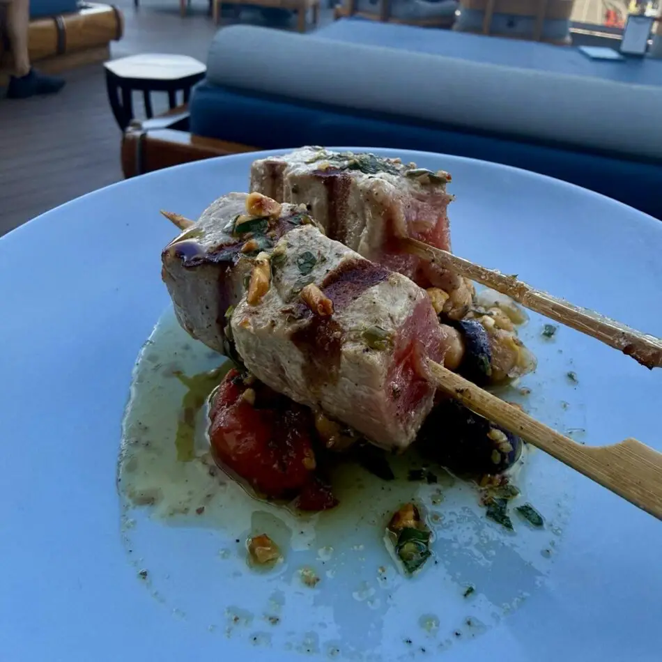 Skewered grilled meat with herbs on a blue plate aboard a Virgin Voyages cruise ship