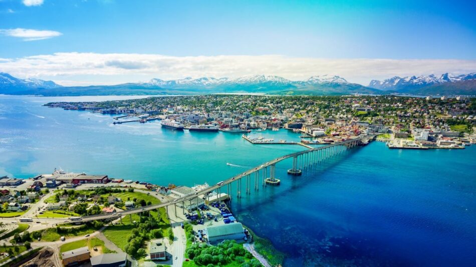 Aerial view of a coastal city with a bridge over blue water, surrounded by mountains under a clear sky.