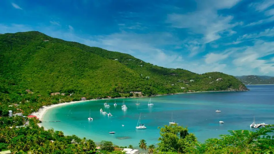 A scenic view of a lush green hillside overlooking a turquoise bay with sailboats and a sandy beach.