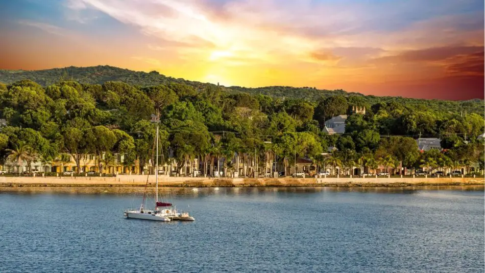 A tranquil bay with a sailboat, lush green trees along the shore, and a vibrant sunset sky.