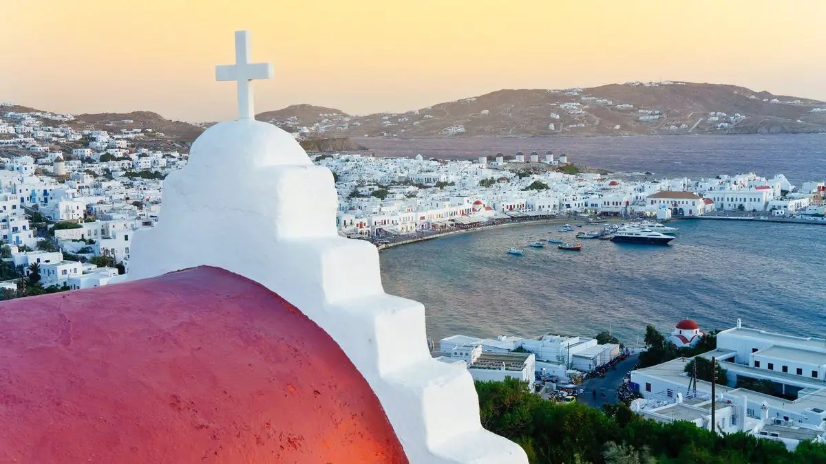 White church dome with a cross overlooks a coastal town and bay at sunset.