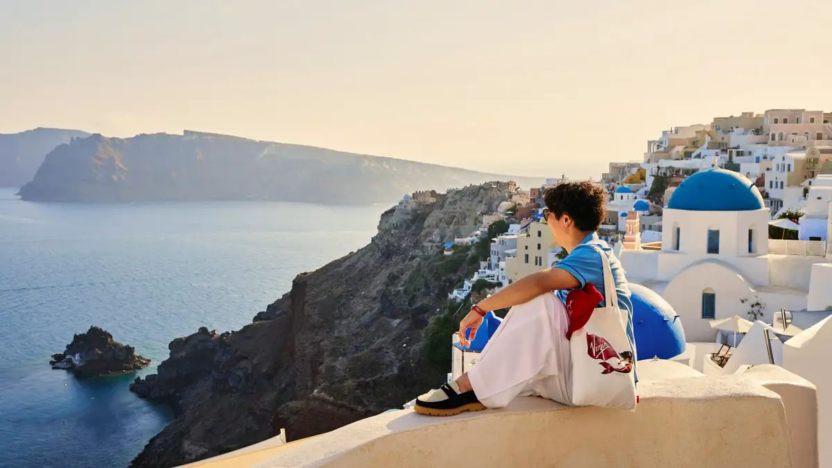 Person sitting on a wall with a view of white buildings and a blue-domed church overlooking the sea and cliffs.