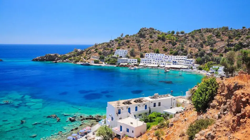 Coastal village with white buildings overlooking a turquoise bay and rugged hills.