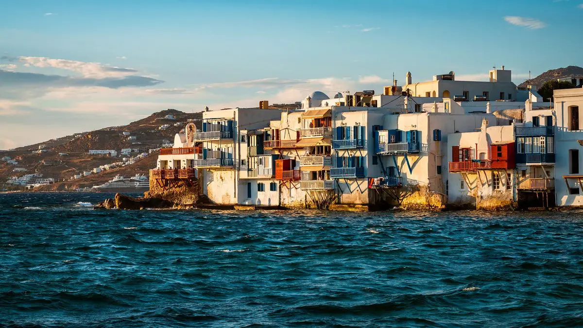 White and blue seaside buildings with balconies overlook the ocean under a partially cloudy sky.