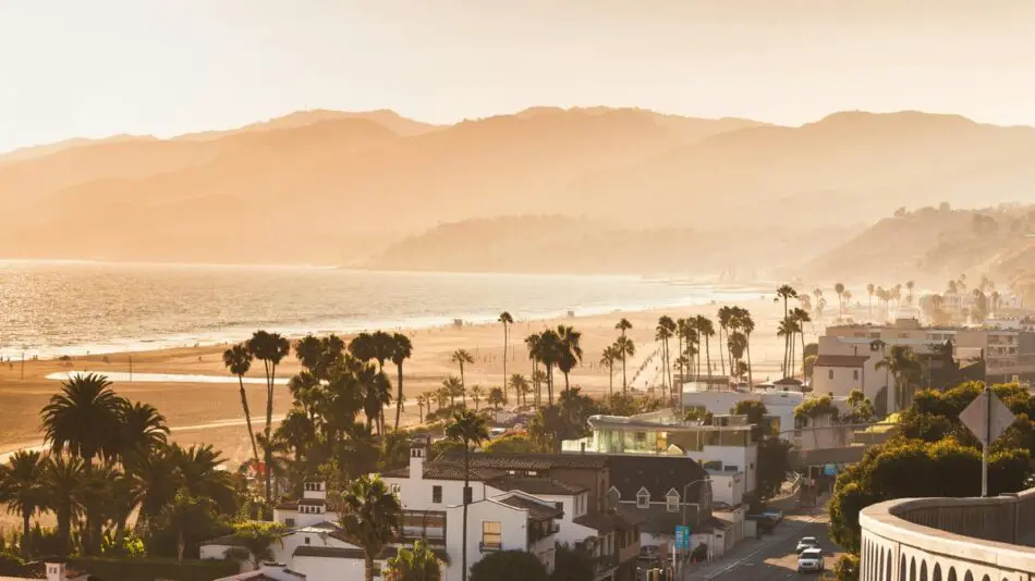 Coastal view with palm trees, a sandy beach, and mountains in the hazy distance.