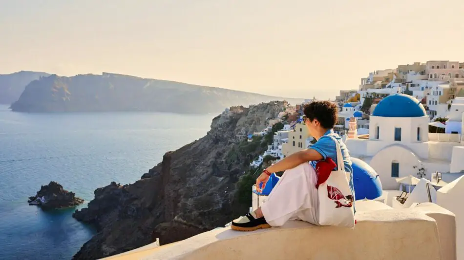 A person sits on a ledge overlooking a seaside village with white buildings and blue domes, against a backdrop of cliffs and a calm sea.