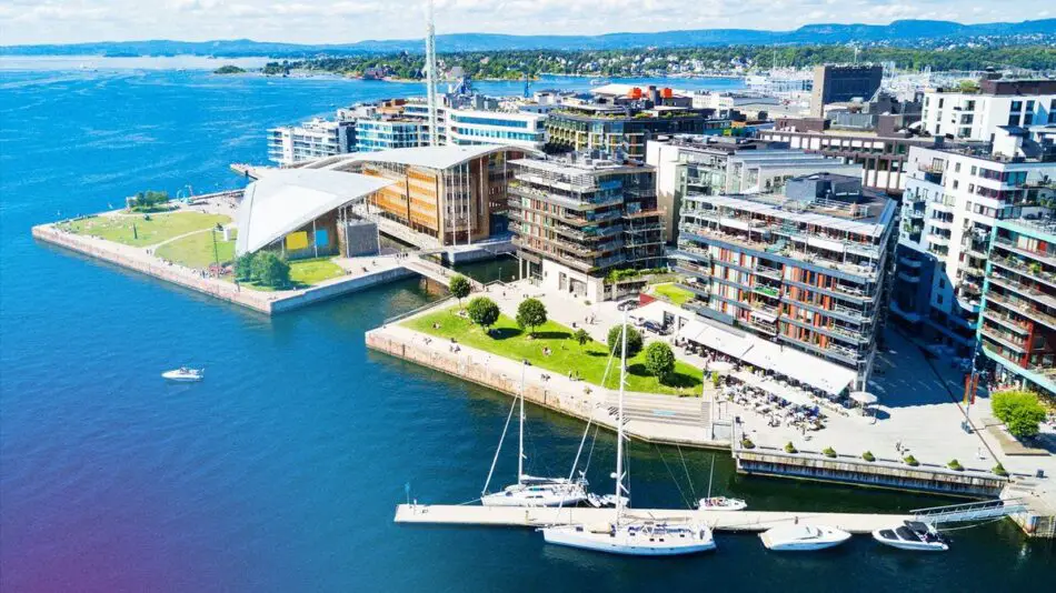 Aerial view of a coastal cityscape with modern buildings, green spaces, and boats docked at a marina.