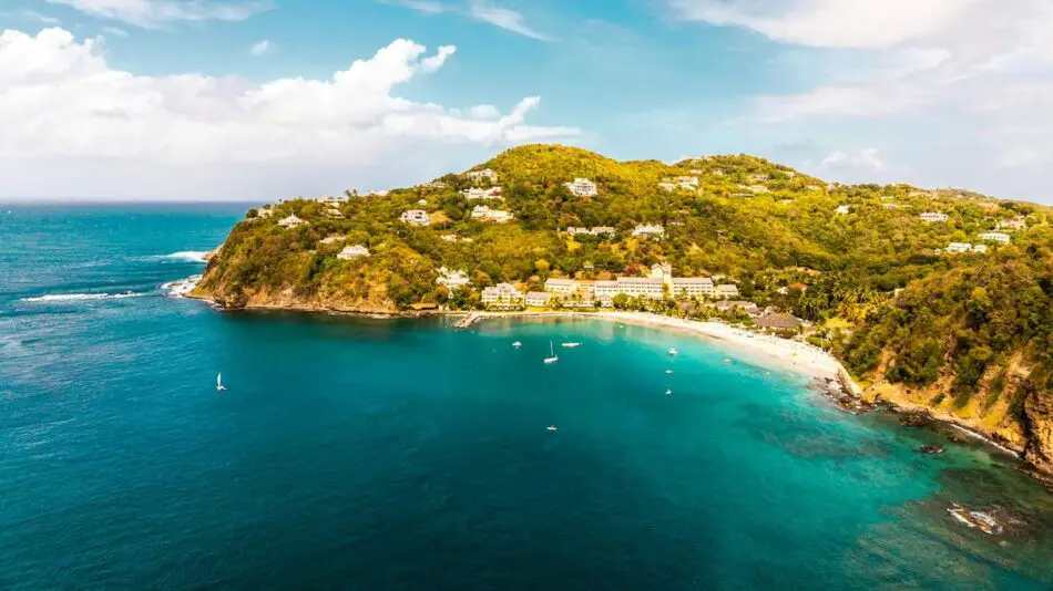 Aerial view of a bay with turquoise waters, sandy beach, lush green hills, and scattered buildings.