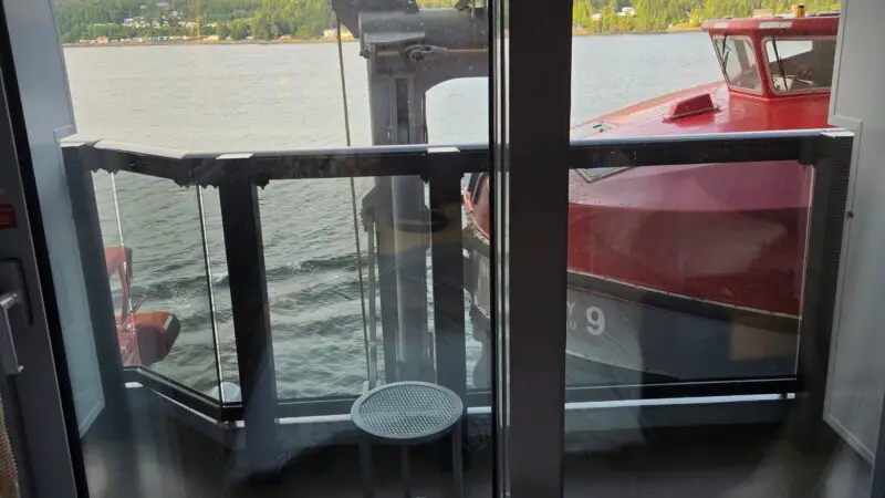 View from a Virgin Voyages cruise ship cabin showing a balcony with a stool, overlooking the sea and distant green hills.