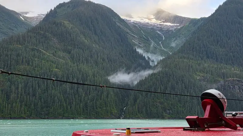 A red Virgin Voyages ship with lush green mountains and a glacier in the background, under a cloudy sky.