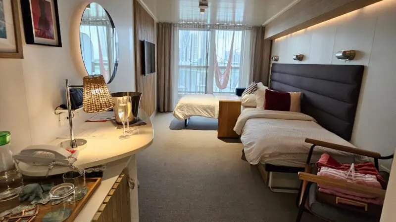 Bright Virgin Voyages cruise ship cabin with a bed, wall-mounted TV, round mirror, desk area, and window with curtains.