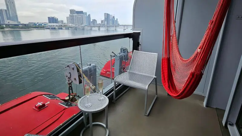 Balcony of a Virgin Voyages cruise ship cabin featuring a red hammock, two chairs, and a small table, overlooking water and a city skyline.