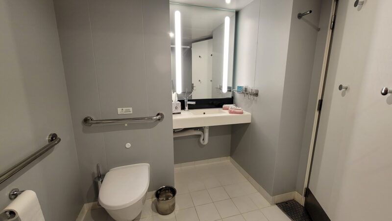 Accessible bathroom in a Virgin Voyages cruise ship cabin, featuring a wall-mounted toilet, safety grab bars, a sink with an illuminated mirror, and towel storage.