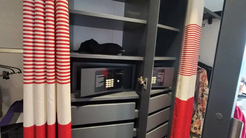 Open closet in a Virgin Voyages cruise ship cabin, featuring life jackets, a safe, and drawers, with red and white striped curtains.