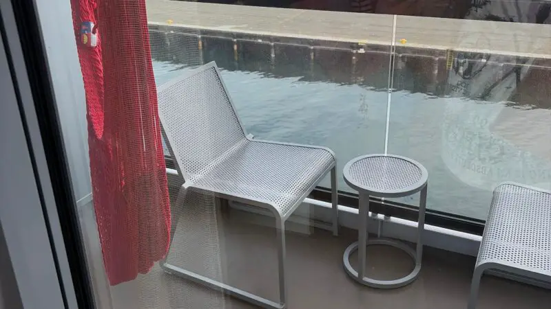 Cruise ship cabin balcony with two chairs, small table, and a view of buildings and greenery through glass railing.