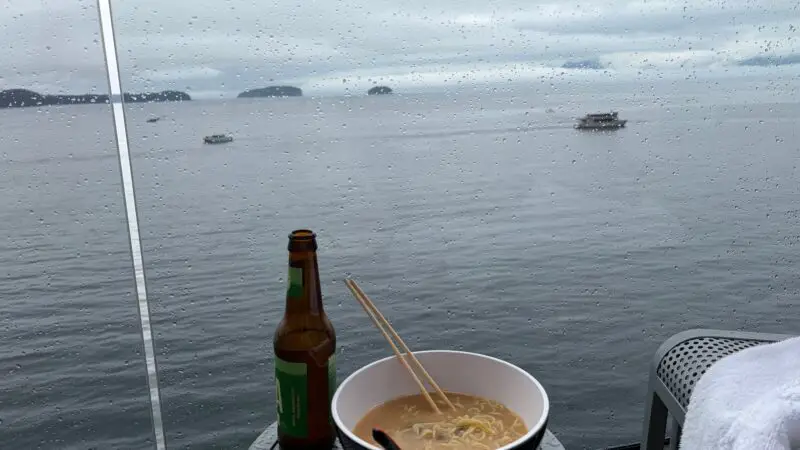 View from a Virgin Voyages cruise ship balcony showing a table with a bowl of noodles and a beer bottle, overlooking a rainy seascape with distant landmasses.