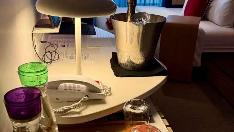 Virgin Voyages cruise ship cabin with a champagne bucket, bottles, glasses, and a phone on a table; bed and balcony with curtains in the background.