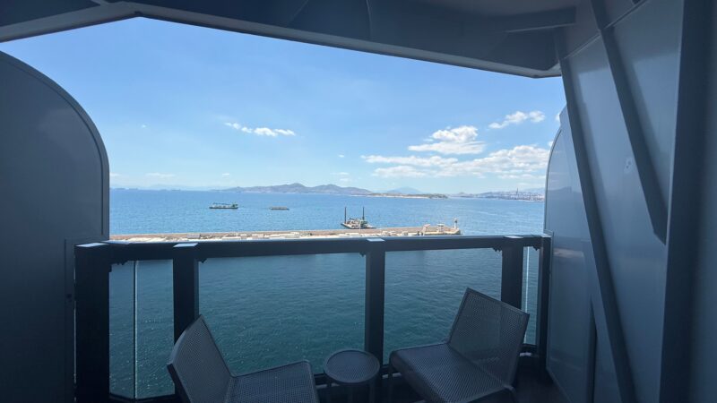 View from a Virgin Voyages cruise ship balcony cabin featuring two chairs and a table, overlooking the ocean and distant coastline.