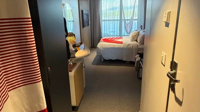 View of a Virgin Voyages cruise ship cabin with a bed, an open balcony curtain, and a hammock visible outside.