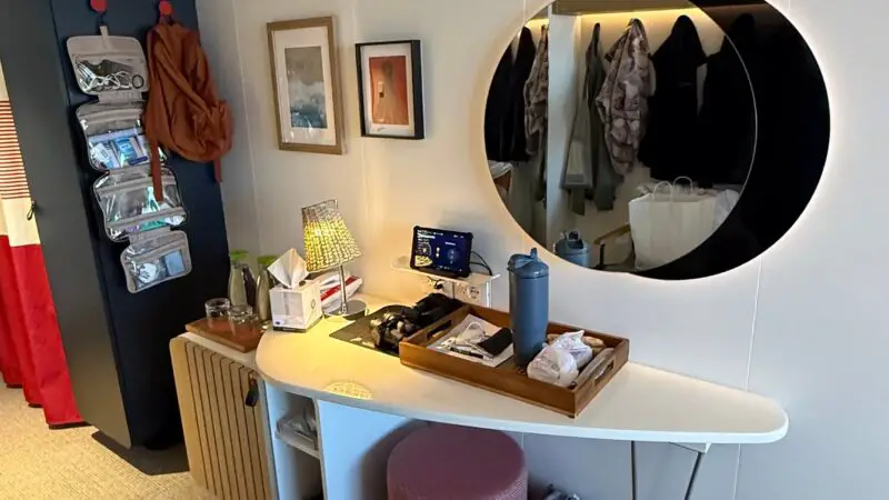 Interior of a Virgin Voyages cruise ship cabin featuring a vanity table with a round mirror, a pink stool, and wall hooks with hanging items.