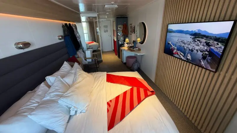 Interior of a Virgin Voyages cruise ship cabin featuring a bed with red and white linens, a wall-mounted TV, and a small desk with a mirror.