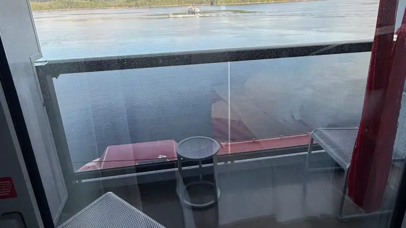 View from a cruise ship cabin showing a balcony with seating, overlooking calm water and forested hills under a blue, cloudy sky.