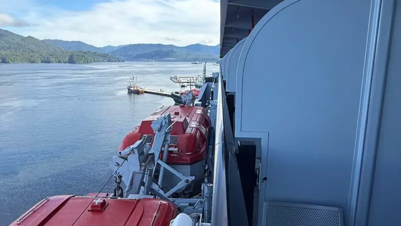 View from a Virgin Voyages cruise ship balcony, showing a metal chair and red lifeboats against a backdrop of calm water and distant mountains.