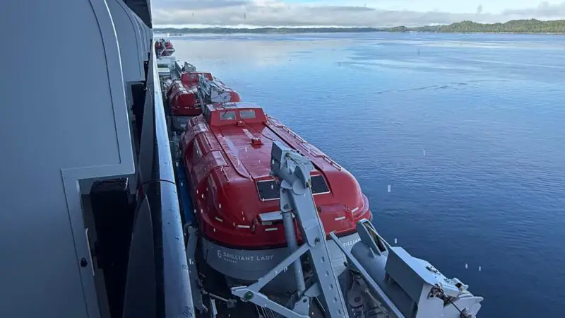 View from a Virgin Voyages cruise ship balcony showing the ocean and red lifeboats along the side of the ship. A hand rests on the railing.