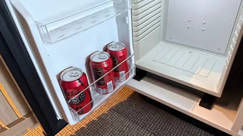 Open mini-fridge in a Virgin Voyages cruise ship cabin, showing three cans of soda on the door shelf.